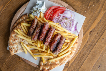 A casual plate featuring grilled kofte, golden french fries, warm pita bread, fresh tomato, red onion, and creamy tzatziki, served on a wooden board.