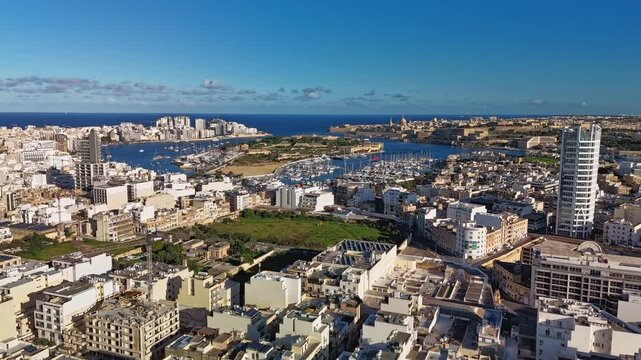Aerial timelapse of Malta's urban skyline, showcasing Ta' Xbiex, Valletta, Sliema, Manoel Island, marina, and the Mediterranean Sea framed by a mix of modern architecture under a sunny winter sky.