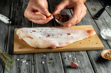 A person sprinkles spices over a fish fillet resting on a wooden cutting board. The kitchen is well-lit with various ingredients and utensils nearby
