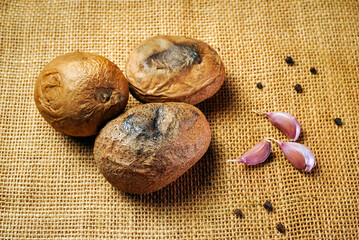 Three potatoes sit on burlap fabric next to two garlic cloves and some black pepper. The scene shows a simple kitchen setup with natural ingredients