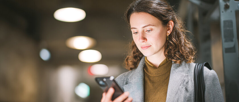 A woman with curly hair checks her smartphone while standing in a well-lit indoor space, showcasing a modern and focused demeanor. - Powered by Adobe