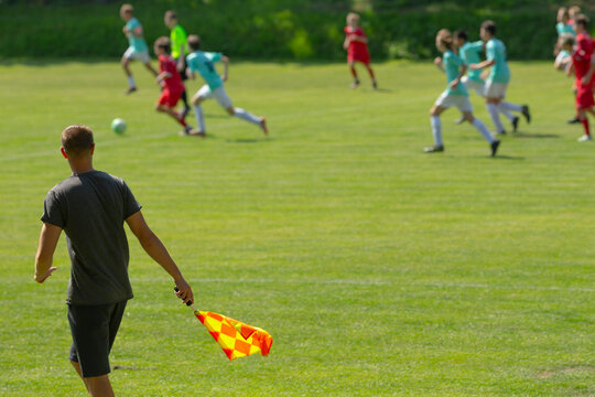 assistant referee holding flag on sideline overseeing youth soccer match in bright sunlight, players in teal and red kits sprinting