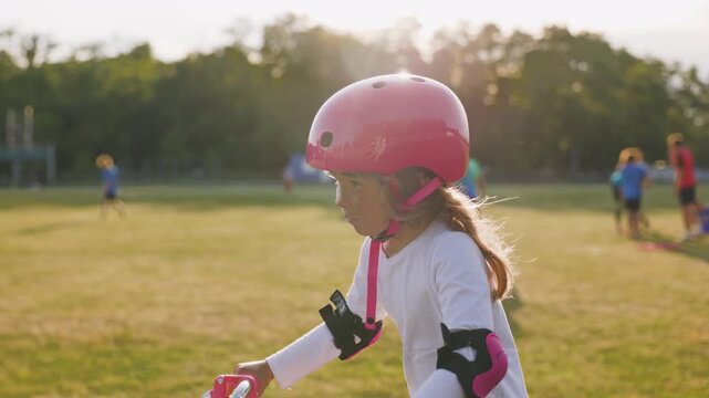 Little girl wearing pink helmet and protective pads learning to ride a bicycle outdoors near a sports field on a sunny summer day