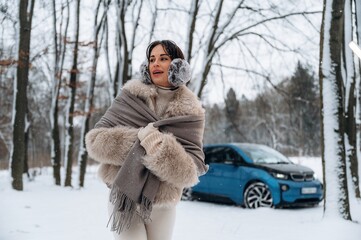 Woman in winter attire, wrapped in a cozy fur shawl, enjoys a snowy landscape with trees and a blue electric car parked nearby, embodying winter fashion and lifestyle