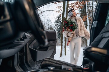 Woman in winter attire holding festive bouquet stands outside car in snowy landscape, showcasing holiday spirit and joy in a serene outdoor environment with nature