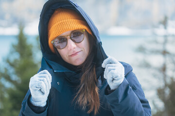 Young woman adjusting her dark blue parka hood over an orange knit hat and sunglasses in snowy mountains