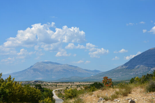 A narrow asphalt road winds through the dry vegetation east of the Peruca Lake with the distant peaks of the Dinara mountain range visible on the horizon on the North
