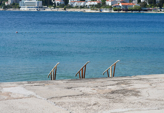 Three rusty metal handrails of sea access ladders stand on a concrete promenade in Selce overlooking the blue Adriatic Sea and coastal architecture.