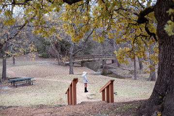 Asian lady in white coat and red skirt stands near wooden bridge, surrounded by leaf-covered paths, picnic tables, and sparse trees, evoking solitude and seasonal beauty, Coppell, Dallas, Texas