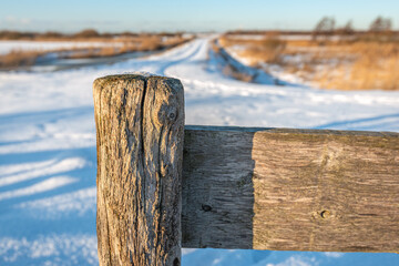 Weathered post of a wooden gate in the foreground of a rural winter landscape with snow.