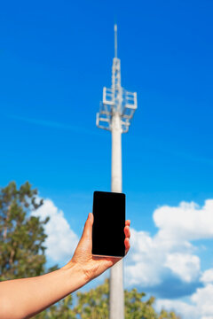A person's hand holds a black smartphone against a blurred background of a tall telecommunication tower and a bright blue sky with white clouds.
