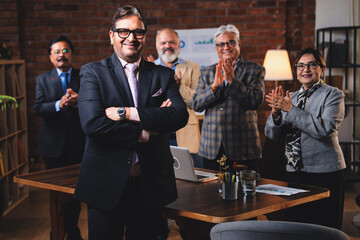 Senior Indian male executive smiling at camera with colleague standing in modern office