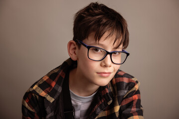 Fototapeta premium Thoughtful studio portrait of a young boy wearing eyeglasses and a casual plaid shirt, neutral beige background, soft natural light