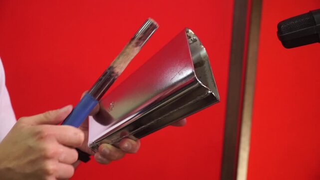 Close-up of a musician playing a metal cowbell with a wooden drumstick against a vibrant red background.