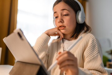 Concentrated child girl studies with tablet and headphones at a desk at home.
