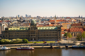 Obraz premium A panoramic view of Prague's riverfront featuring a grand historic building with a green dome, red tiled rooftops, and bustling boats along the river, capturing Central European charm and architecture