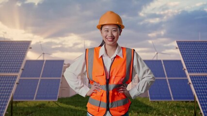 Asian Female Engineer Wearing Safety Helmet Smiling To Camera While Standing With Arms Akimbo at Energy Farm featuring Solar Panels, Battery Storage and Wind Turbines