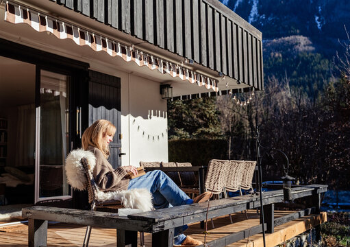 Woman journaling and reflecting on new year intentions while relaxing on wooden sunny terrace of chalet with mountain view. Mindfulness, slow living, wellbeing, self care, mental health concept
