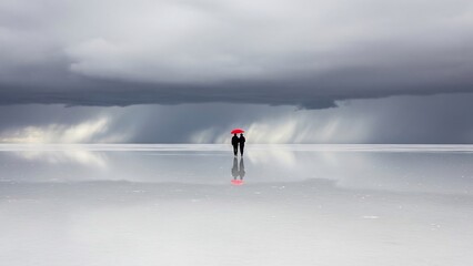 Romantic couple with a vibrant red umbrella walking on a reflective salt flat under a dramatic stormy sky with distant rain. Ideal for travel blogs, anniversary cards, inspirational content,