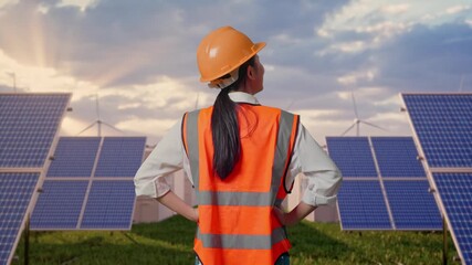 Back View Of A Female Engineer Wearing Safety Helmet Looking Around While Standing With Arms Akimbo at Energy Farm featuring Solar Panels, Battery Storage and Wind Turbines