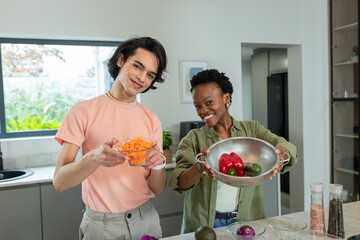 Diverse friends standing at marble island holding bowls of carrots peppers avocados and onion