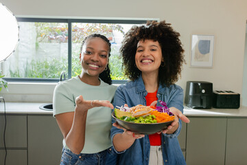 Diverse female friends holding colorful salad bowl in modern kitchen with ring light and countertop