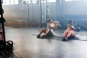 Two mid-adult female exercisers holding medicine balls and twisting on mats in gym, copy space