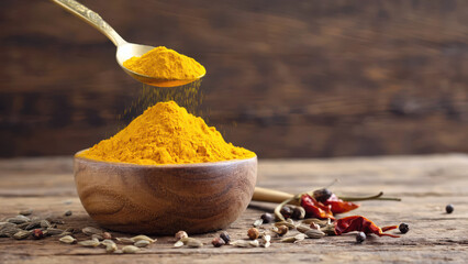 A wooden bowl holds bright turmeric powder while a brass spoon pours fine grains. Cumin seeds and dried chilies are scattered around on a rustic kitchen table
