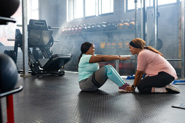 Diverse female workout partners performing sit-up on rubber gym mat holding ankles near kettlebells