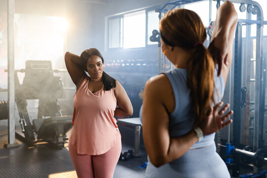 Diverse female partners in pink sportswear stretching and guiding at gym by mirror and weights