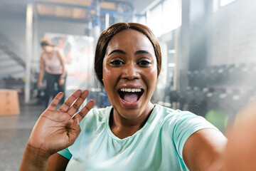 Diverse female friends waving and smiling while training near dumbbell rack in turquoise top