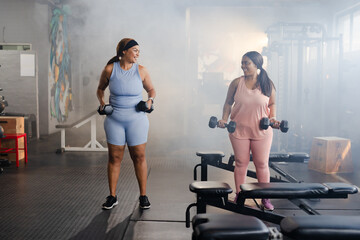 Diverse female friends in athletic clothing lifting hex dumbbells while smiling in gym weight area