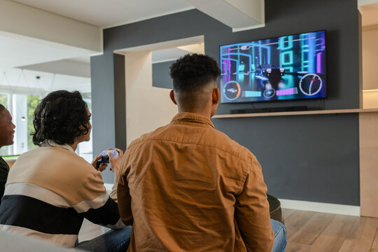 Diverse men sitting on sofa in living room playing racing game on TV with white controller