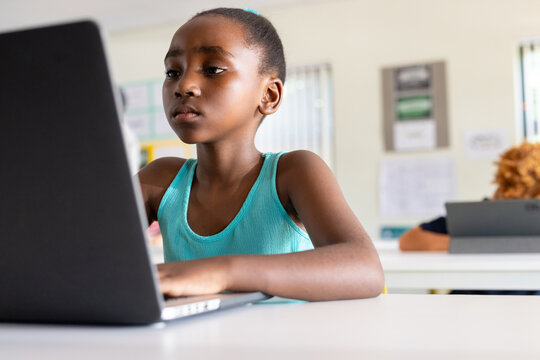 African female child sitting at desk in classroom using laptop, wearing teal sleeveless top