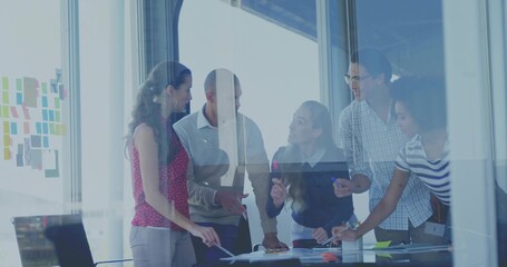 Leaning over conference table, five colleagues pointing at papers in glass room with sticky notes