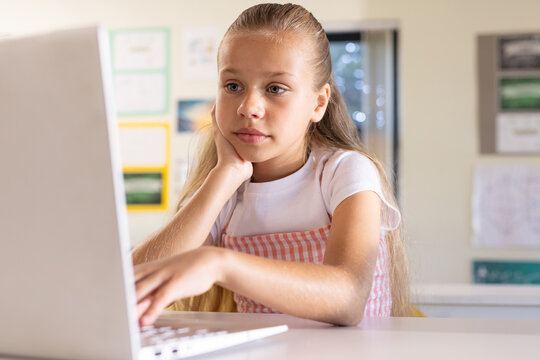 Female child sitting in school working on laptop, resting chin, wearing pink gingham dress