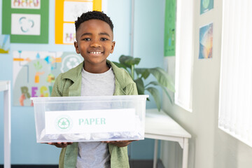 African American youth boy holding labeled PAPER bin in classroom recycling corner, smiling