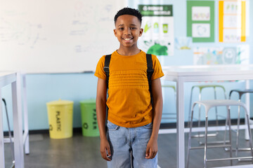 African American boy standing in classroom by lab tables wearing mustard tee and backpack near bins