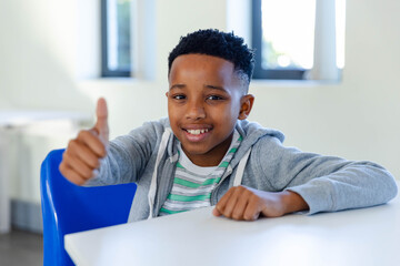African American youth male giving thumbs-up while sitting at white table in classroom in hoodie