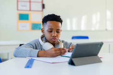 African American boy studying at desk in class wearing gray sweatshirt and headphones using tablet