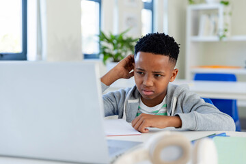 African adolescent male student sitting at classroom desk using laptop, notebook, pen, potted plant