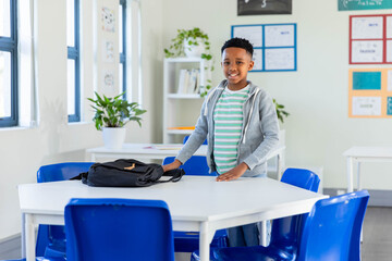 Youth male student standing at white table in classroom, holding black backpack near window