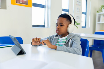 African American teenage boy wearing hoodie, white headset, tapping tablet on stand at school desk