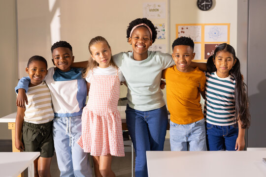 Diverse group of children standing with arms around each other, smiling in classroom near desks