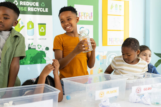 Diverse children gathering at school table holding glass jar and sorting PLASTIC and GLASS bins