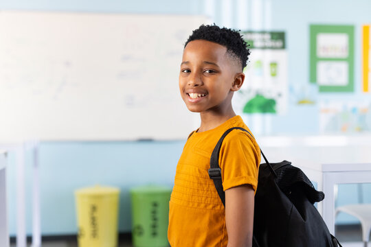African-American child standing in classroom with black backpack near recycling bins, copy space