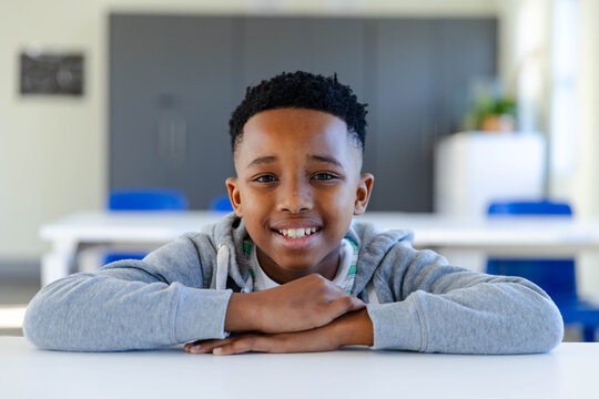 African American child boy sitting at white classroom table, leaning on arms, wearing gray hoodie