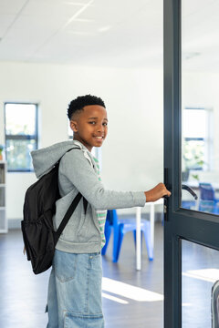 African-American teen male with black backpack standing at glass door holding metal handle smiling