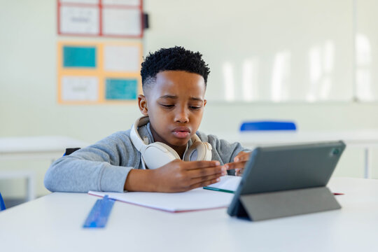 African American boy studying at desk in class wearing gray sweatshirt and headphones using tablet