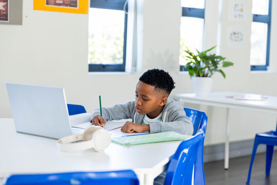 African male youth sitting at classroom table writing with pencil in notebook beside open laptop